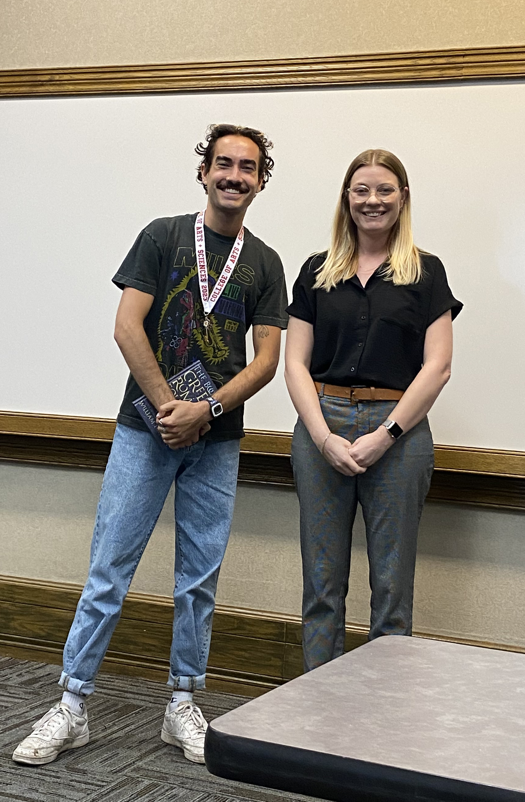 Professor and graduating senior holding a book and wearing a lanyard, smiling at the camera